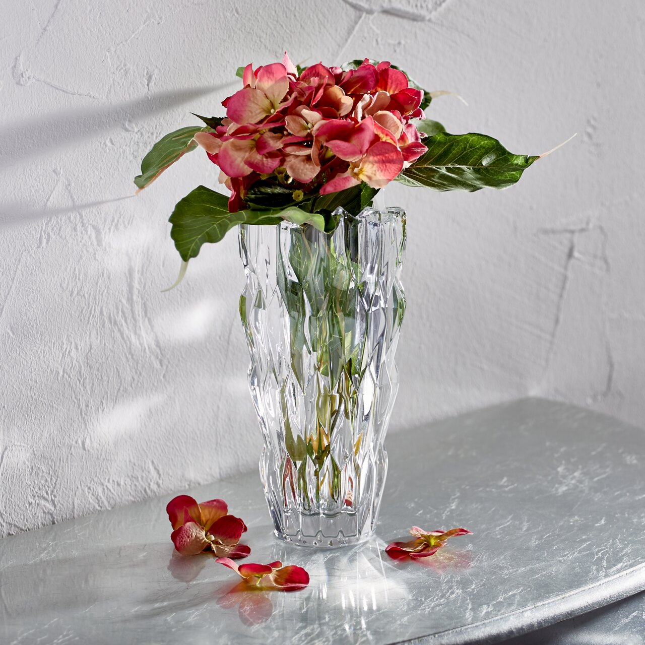 Kristallvase mit facettierter Oberfläche, gefüllt mit rosa Blumen, auf einem Tisch vor einer strukturierten Wand, seitliche Perspektive