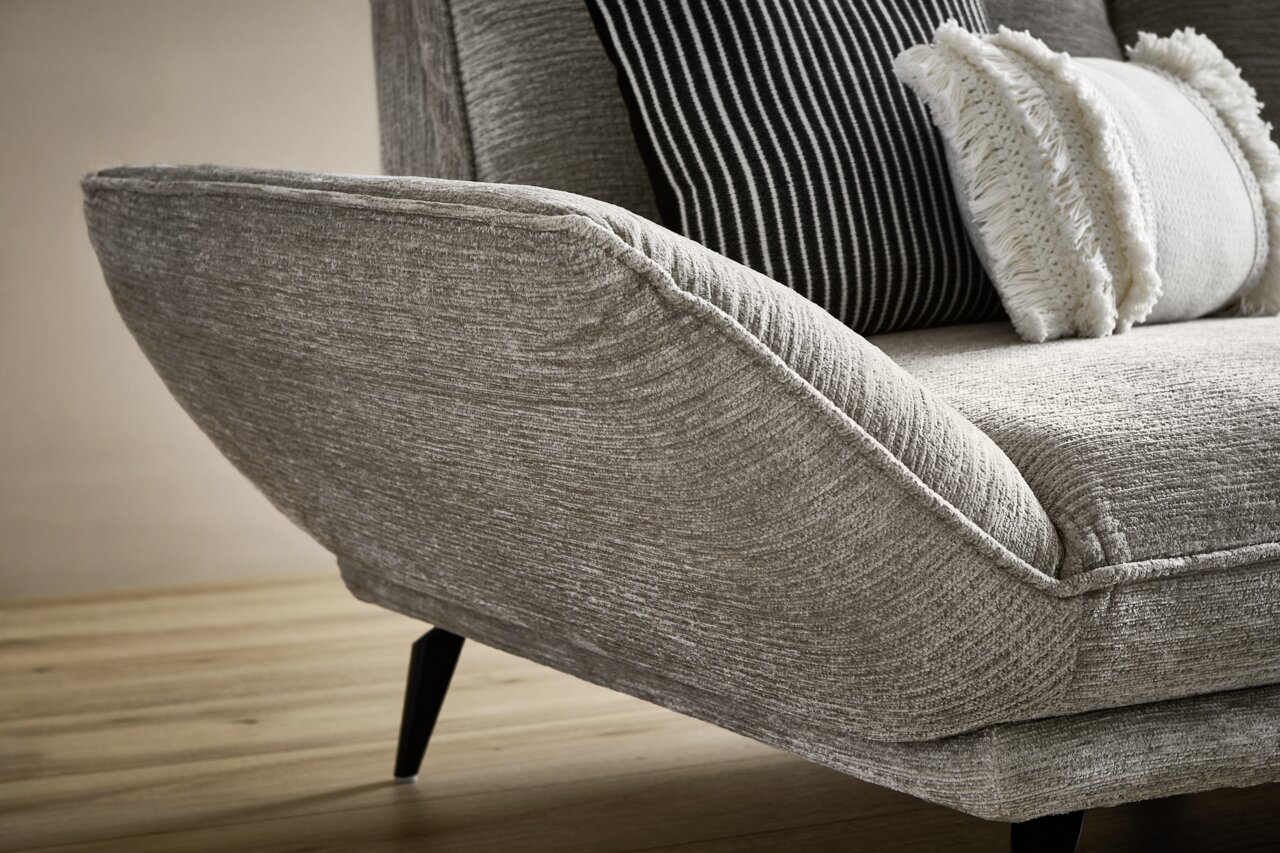 Nahaufnahme eines modernen Longchairs mit grauem Stoffbezug und schwarzen, schrägen Beinen. Im Hintergrund sind dekorative Kissen zu sehen. Perspektive von der Seite.