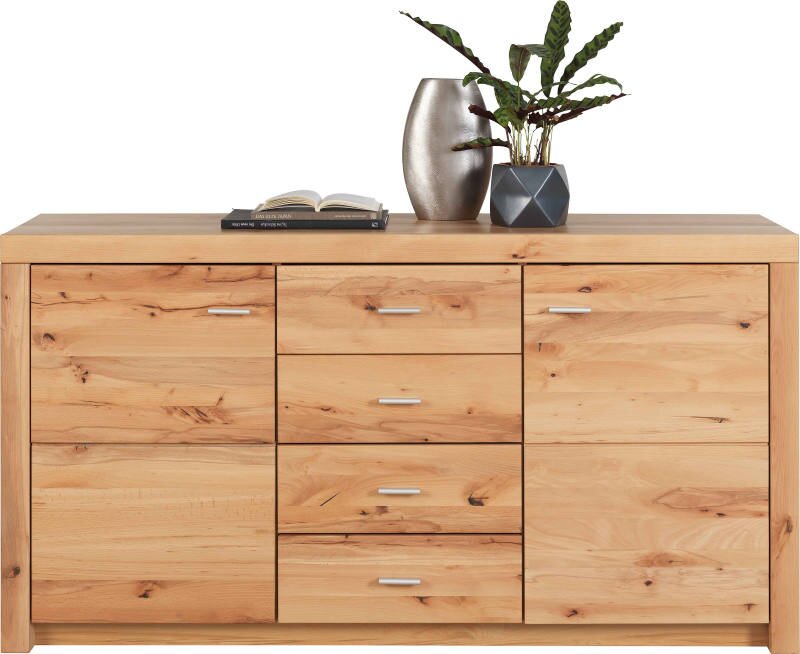 Vooraanzicht van een brede houten dressoir/commode met natuurlijke nerf en knoesten, vier centrale lades met metalen grepen en twee zijdeuren, met vaas, boek en plant bovenop tegen witte achtergrond