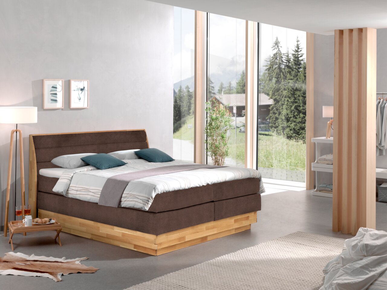 Modernes Boxspringbett in einem hellen, stilvollen Schlafzimmer mit großen Fenstern und Blick auf die Natur, seitliche Perspektive.