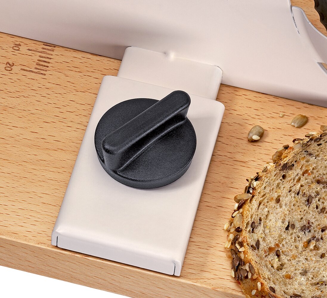 Close-up schuin van boven op een houten snijplank met een wit kunststof accessoire met zwarte draaiknop, naast een snee meergranenbrood.