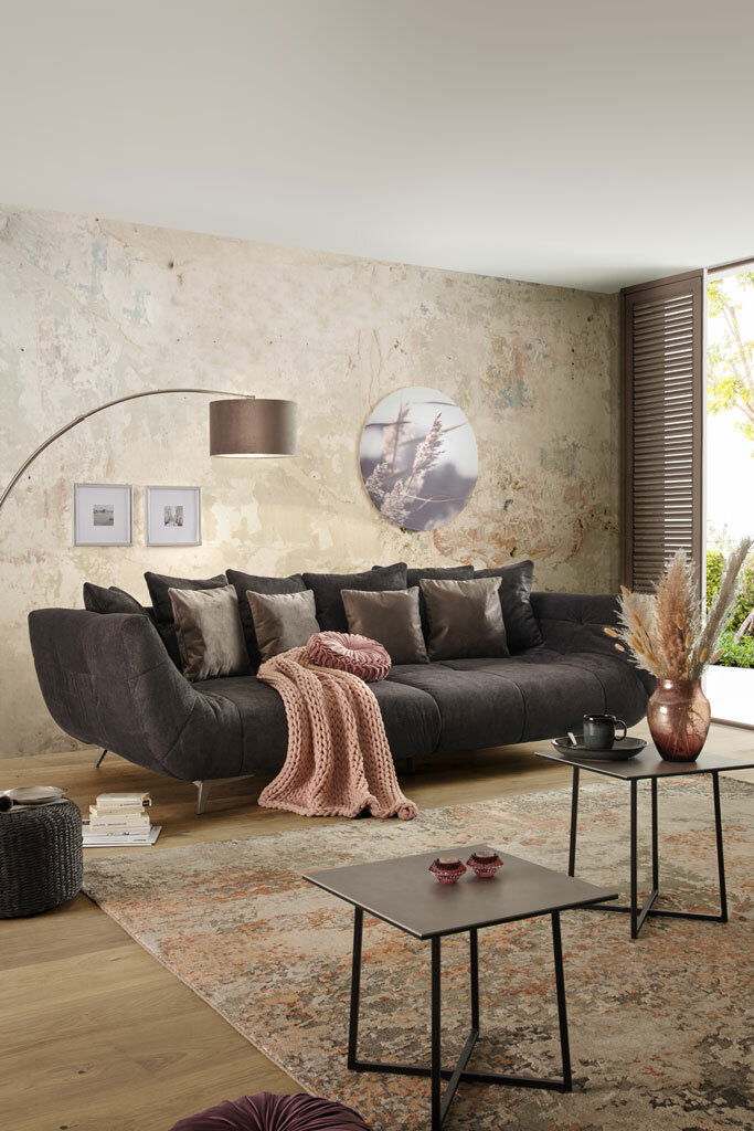 Großes, modernes Sofa in dunklem Grau mit mehreren Kissen, dekoriert mit einer rosa Decke, in einem stilvoll eingerichteten Wohnzimmer. Perspektive von vorne.
