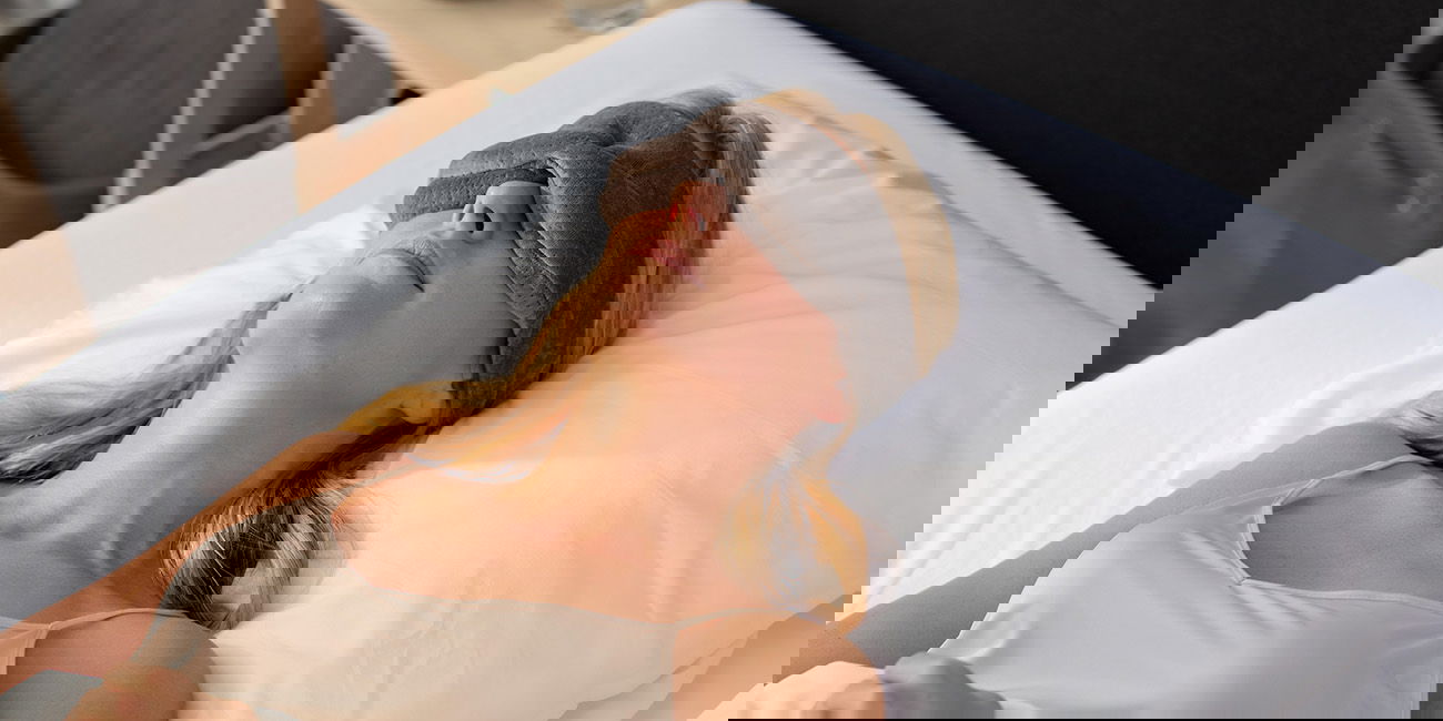 eine Frau mit Schlafmaske liegt im Bett mit weißem Kissen.