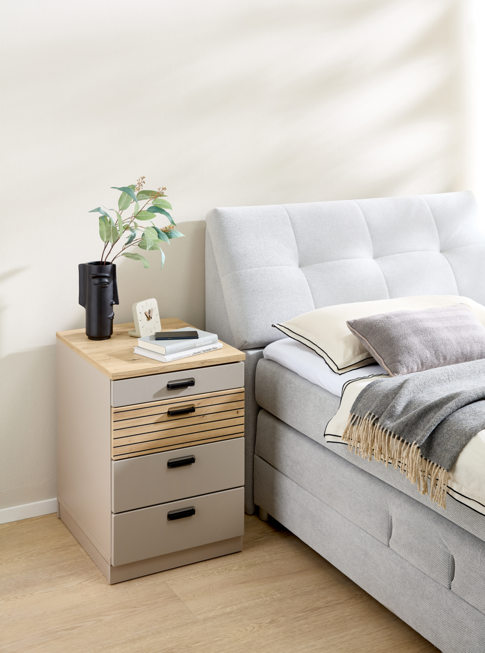 Rollcontainer mit vier Schubladen, aus hellem Holz und Metall, neben einem Bett in einem modernen Schlafzimmer, aus seitlicher Perspektive.