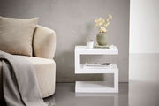 Moderner weißer Beistelltisch in Z-Form, seitlich fotografiert, neben einem beigen Sofa mit Kissen und Decke, dekoriert mit einer Vase, Tasse und Büchern.