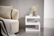Moderner weißer Beistelltisch in Z-Form, seitlich fotografiert, neben einem beigen Sofa mit Kissen und Decke, dekoriert mit einer Vase, Tasse und Büchern.