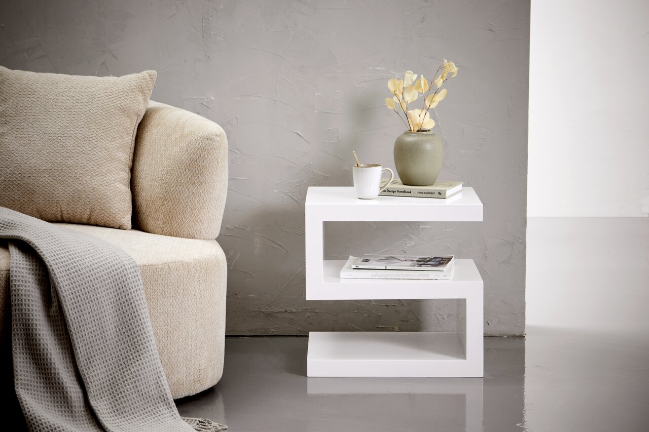 XORA Beistelltisch KENZO Moderner weißer Beistelltisch in Z-Form, seitlich fotografiert, neben einem beigen Sofa mit Kissen und Decke, dekoriert mit einer Vase, Tasse und Büchern.
