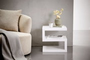 Moderner weißer Beistelltisch in Z-Form, seitlich fotografiert, neben einem beigen Sofa mit Kissen und Decke, dekoriert mit einer Vase, Tasse und Büchern.
