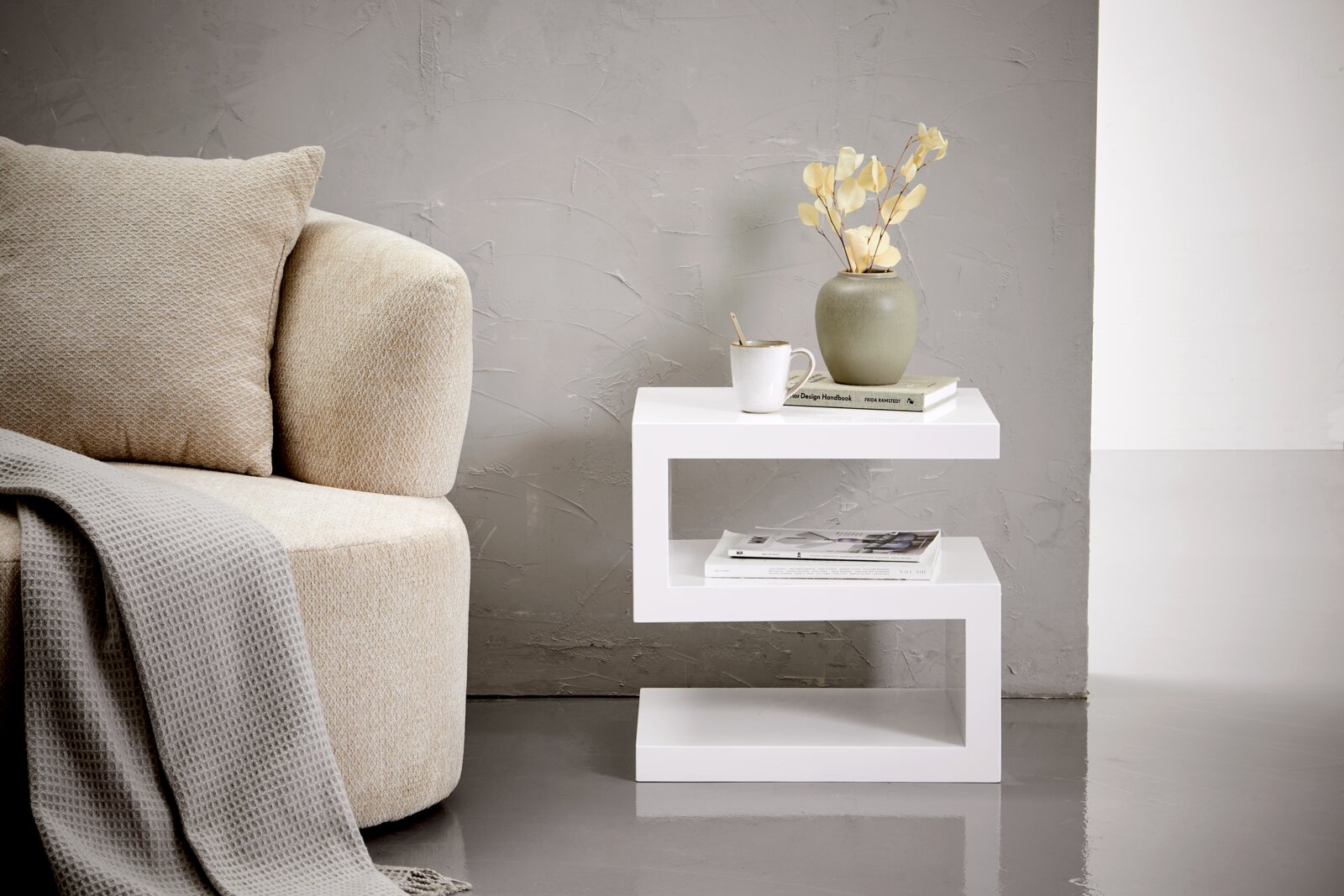 Moderner weißer Beistelltisch in Z-Form, seitlich fotografiert, neben einem beigen Sofa mit Kissen und Decke, dekoriert mit einer Vase, Tasse und Büchern.