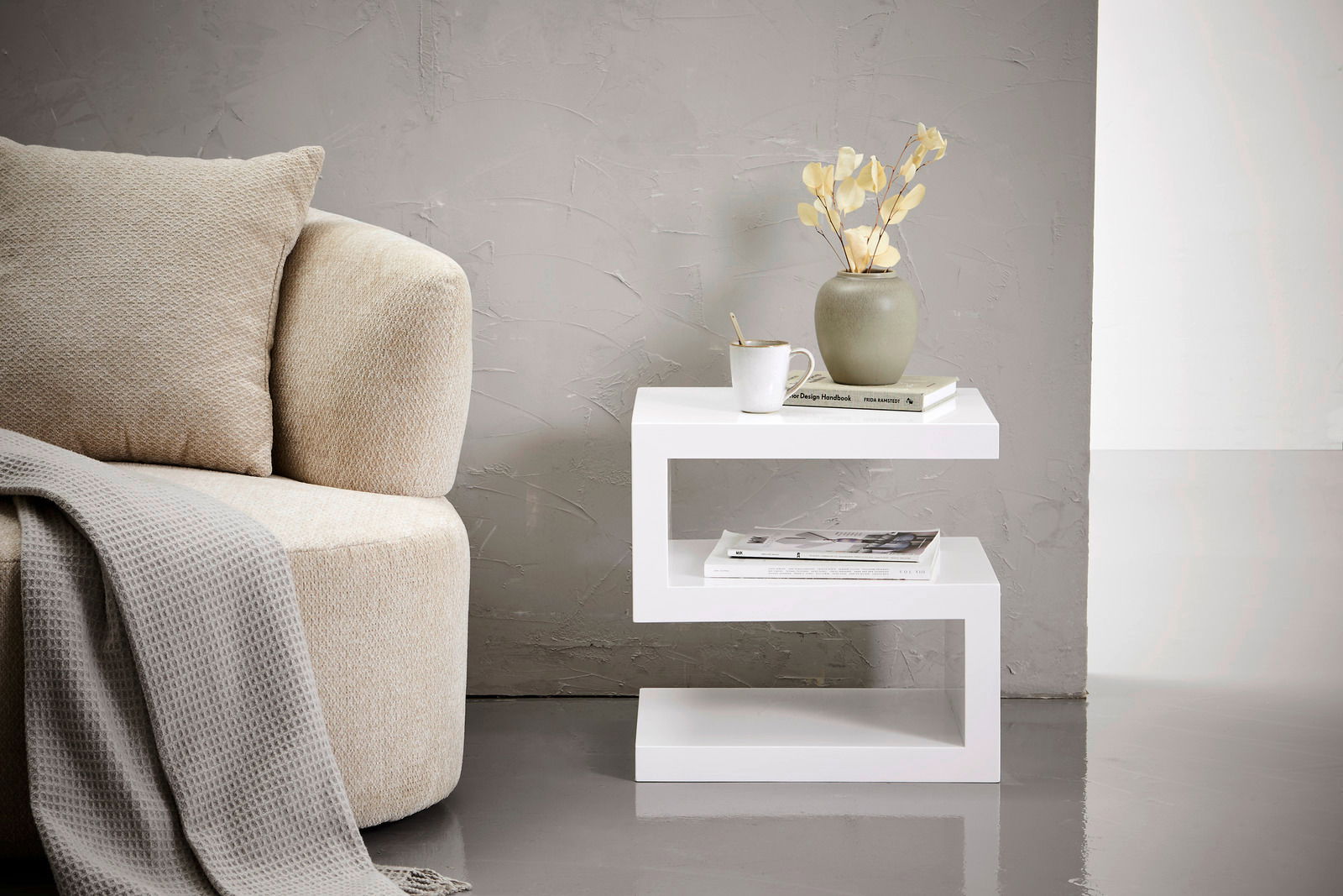Moderner weißer Beistelltisch in Z-Form, seitlich fotografiert, neben einem beigen Sofa mit Kissen und Decke, dekoriert mit einer Vase, Tasse und Büchern.