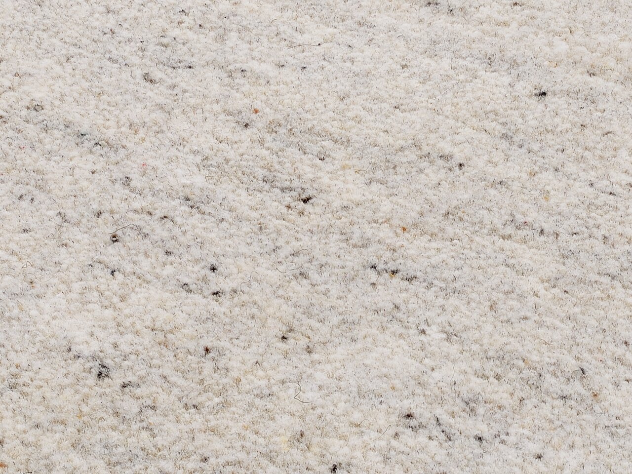 Nahaufnahme eines cremefarbenen Wollteppichs mit feiner Textur und vereinzelten dunklen Fasern, von oben betrachtet.