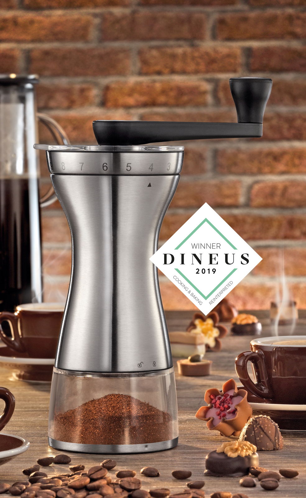 Kaffeemühle Manaos aus Edelstahl mit Kurbel, seitliche Perspektive, umgeben von Kaffeetassen und Gebäck, DINEUS 2019 Gewinner.