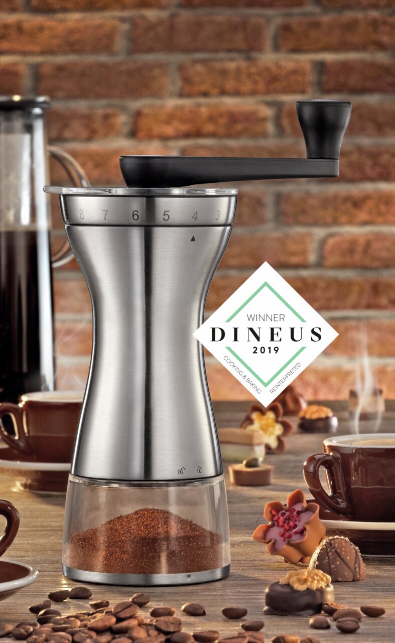 Kaffeemühle Manaos aus Edelstahl mit Kurbel, seitliche Perspektive, umgeben von Kaffeetassen und Gebäck, DINEUS 2019 Gewinner.
