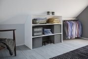 Weißes Ankleide-Regal mit offenen Fächern, gefalteter Kleidung, grauen Aufbewahrungsboxen und Schuhen neben einer Kleiderstange mit Hemden, gezeigt in seitlicher Perspektive von vorn rechts in einem hellen Dachzimmer.