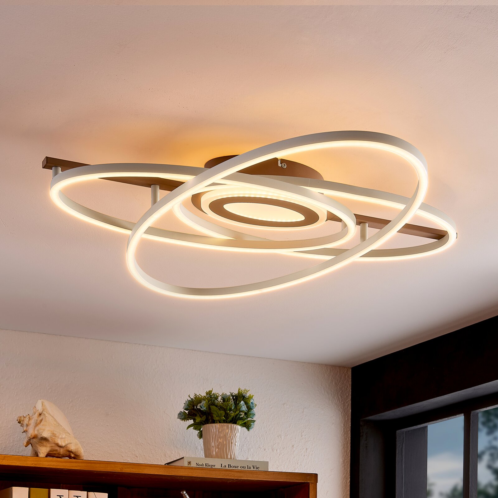 Moderne LED plafondlamp met overlappende ringen in wit met warmwit licht, aan het plafond gemonteerd, gefotografeerd vanuit een lage hoek (onderaanzicht) in een woonkamer.