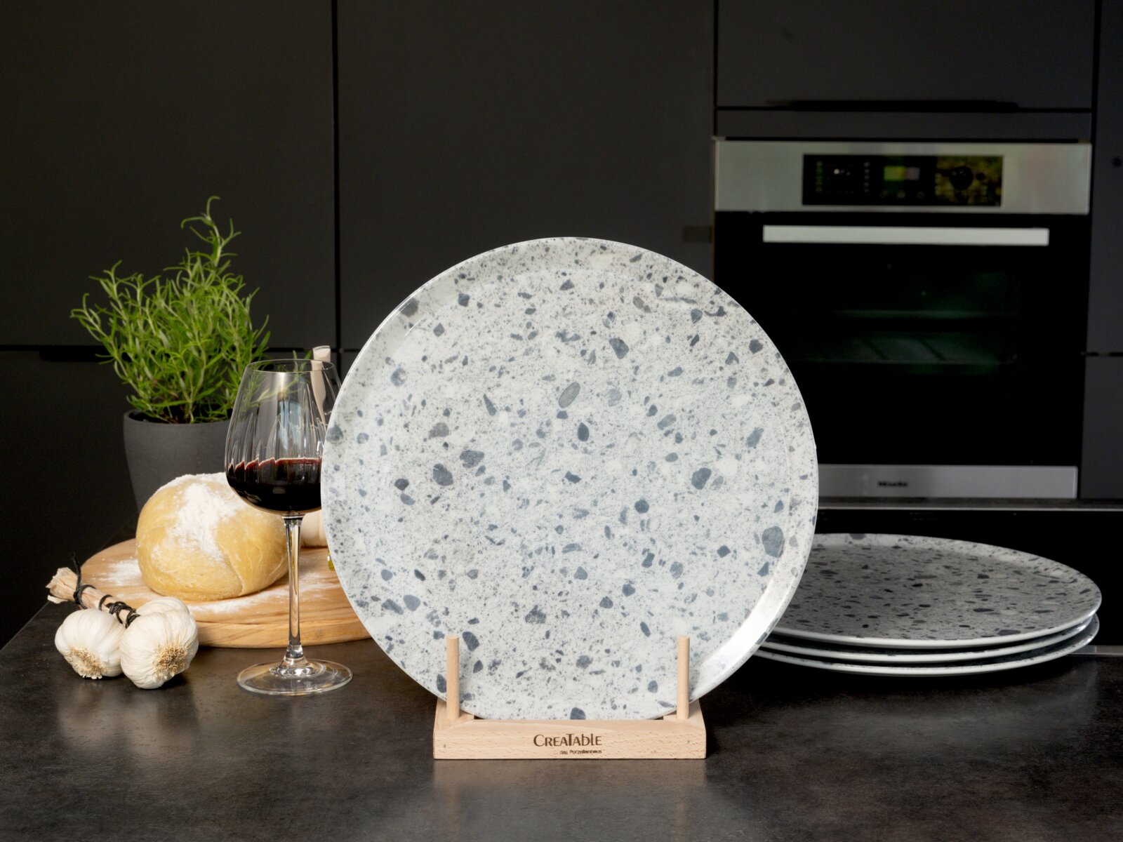 Creatable Pizzateller 4-tlg STONE COLLECTIONF Großer runder Speiseteller in hellgrauer Steinoptik mit dunklen Sprenkeln auf Holzständer, frontal aus Augenhöhe in moderner Küche fotografiert; im Hintergrund weitere Teller, Rotweinglas, Brot, Knoblauch und Kräutertopf.