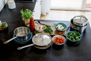 Edelstahl-Kochtopf und Pfanne mit Deckeln auf schwarzer Küchenarbeitsplatte, umgeben von frischen Zutaten wie Spinat, Kirschtomaten, Eiern, Kräutern und Käse; Perspektive: leicht von oben (Top-Down) in schräger Ansicht.