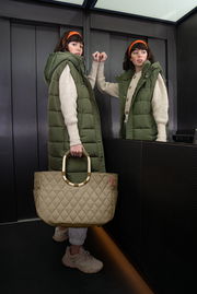 Frau in einem Aufzug mit grünem Steppmantel und beiger, gesteppter Tasche mit runden Griffen, von vorne betrachtet.