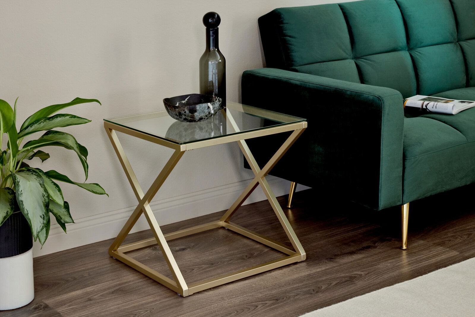 Jahnke Beistell-/Nachttisch X-TRA D 140 Moderner Beistelltisch mit Glasplatte und goldfarbenem Metallgestell, seitliche Perspektive neben einem grünen Sofa.