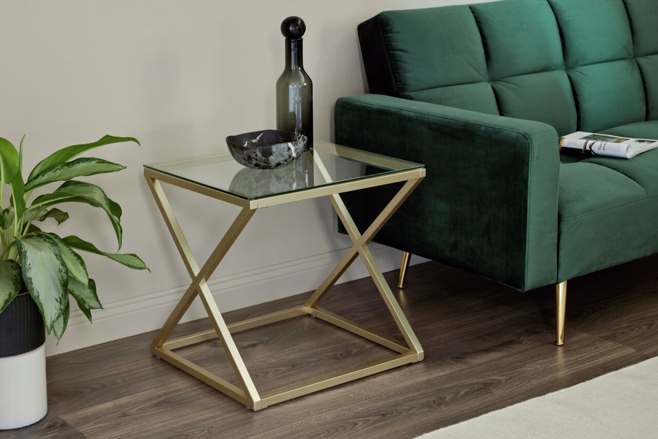 Moderner Beistelltisch mit Glasplatte und goldfarbenem Metallgestell, seitliche Perspektive neben einem grünen Sofa.
