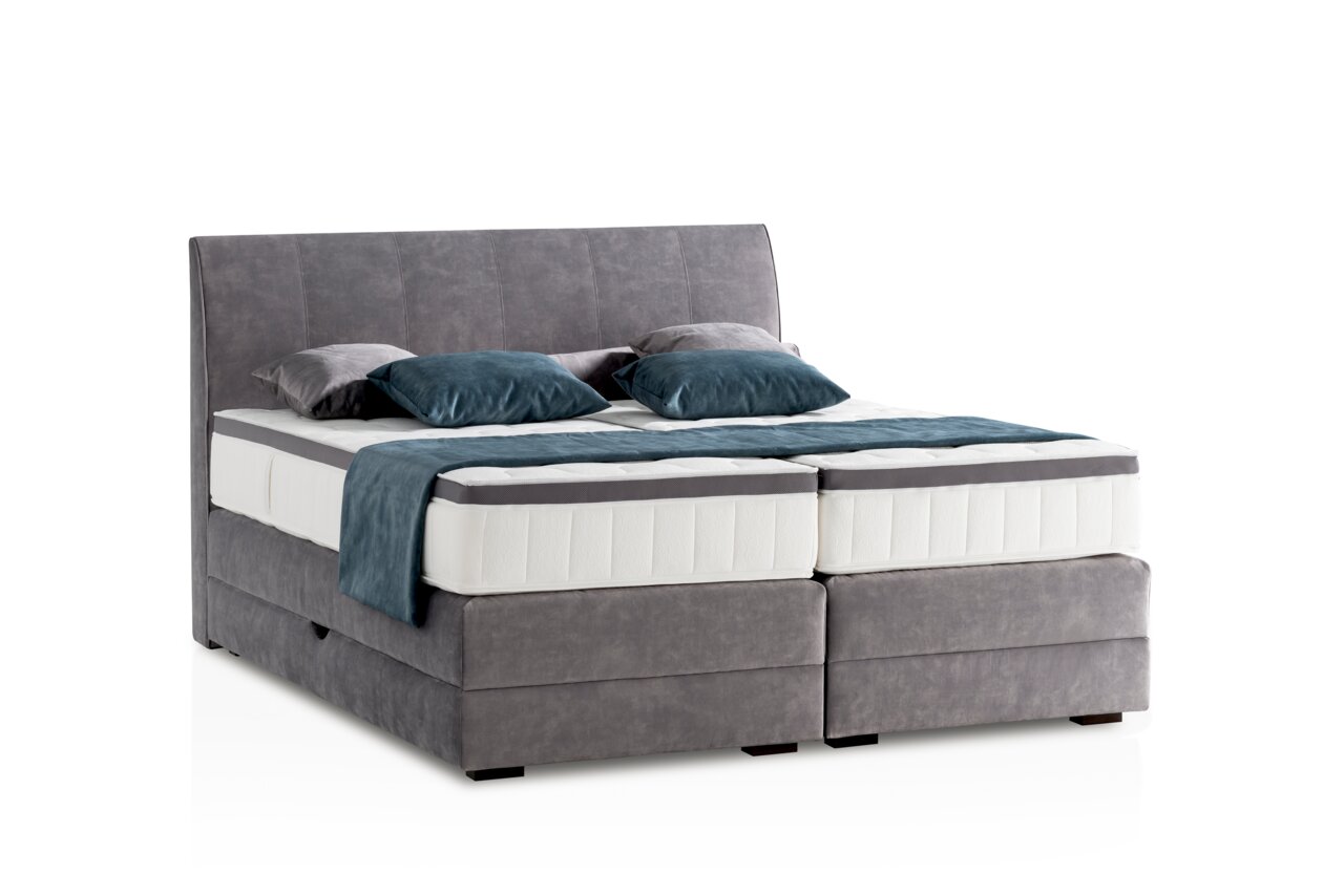 Graues Boxspringbett mit Kopfteil, seitliche Perspektive, dekoriert mit blauen und grauen Kissen.