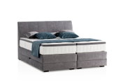Graues Boxspringbett mit Kopfteil, seitliche Perspektive, dekoriert mit blauen und grauen Kissen.