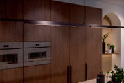 Moderne schwarze lineare LED-Pendelleuchte über einer Kücheninsel vor Holzfronten, seitliche Perspektive in leichtem Untersichtwinkel