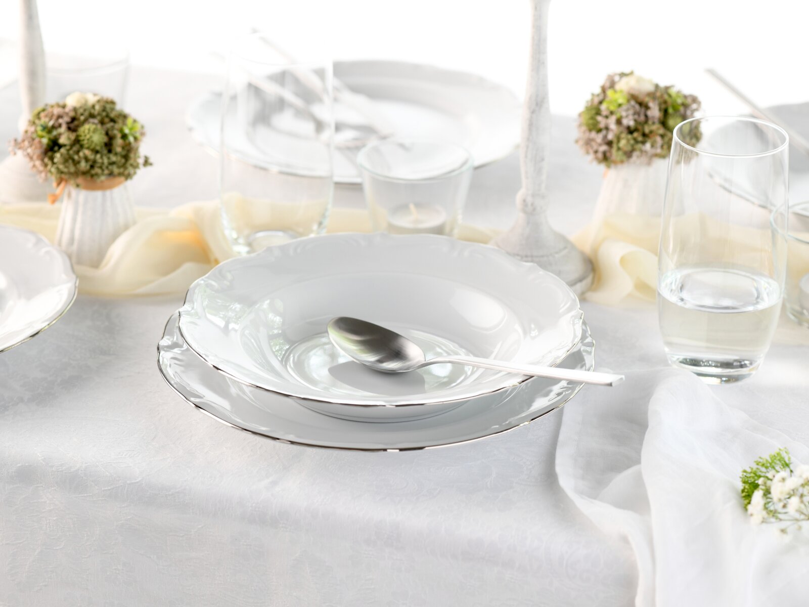 Elegant gedeckter Tisch mit weißem Porzellan-Suppenteller auf Speiseteller mit silbernem Rand, Löffel, Gläsern und Kerze auf weißer Tischdecke, Ansicht schräg von vorn