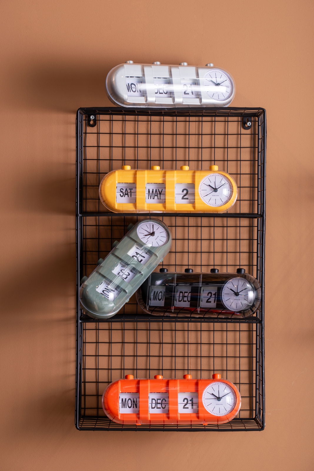 Verschiedene Tischuhren mit Kalenderfunktion in bunten Farben auf einem Gitterregal, seitliche Perspektive.