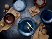 Dunkelblaues Keramikgeschirr-Set mit Tellern und Schalen, goldfarbenem Besteck und Holzbrettchen auf dunklem Tisch, Draufsicht von oben