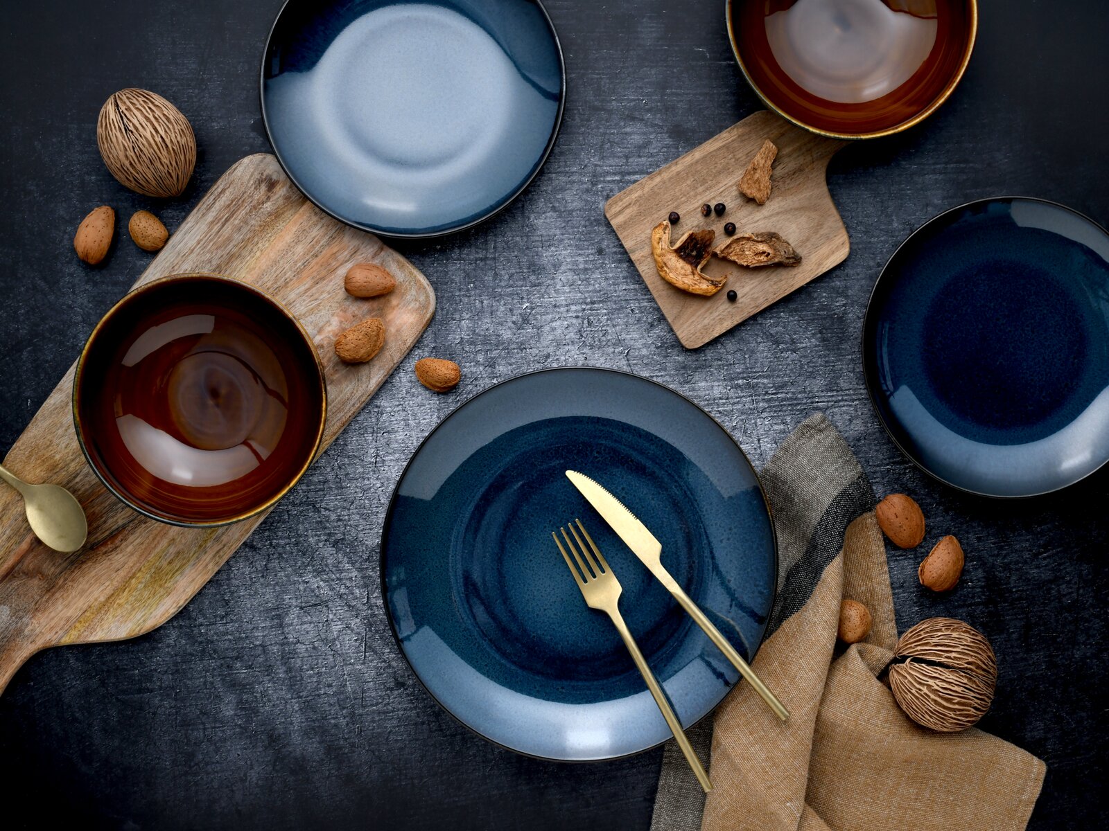 Dunkelblaues Keramikgeschirr-Set mit Tellern und Schalen, goldfarbenem Besteck und Holzbrettchen auf dunklem Tisch, Draufsicht von oben
