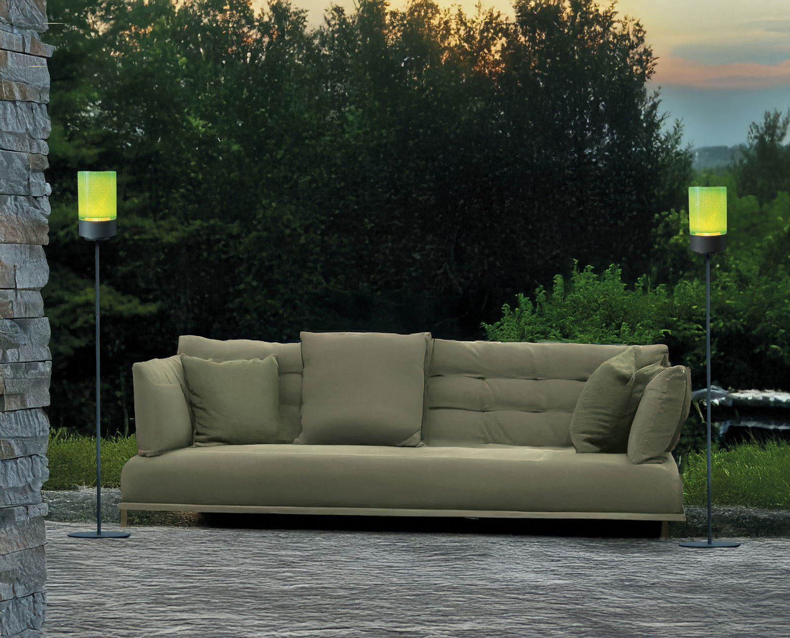 Zwei Akku-Windlichter auf hohen Gestellen flankieren ein grünes Sofa im Freien, umgeben von Natur bei Sonnenuntergang. Perspektive: Frontalansicht.