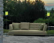 Zwei Akku-Windlichter auf hohen Gestellen flankieren ein grünes Sofa im Freien, umgeben von Natur bei Sonnenuntergang. Perspektive: Frontalansicht.