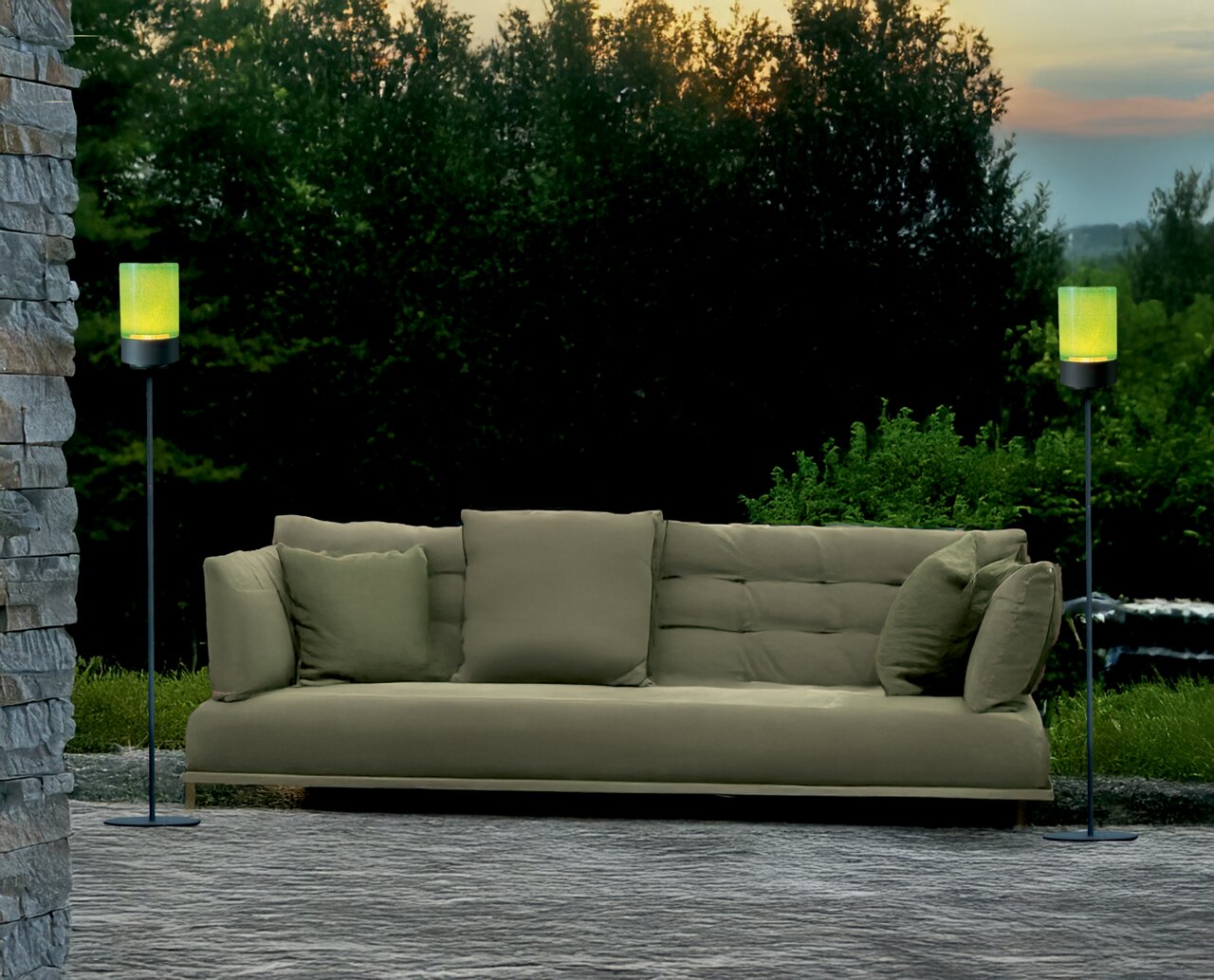 Zwei Akku-Windlichter auf hohen Gestellen flankieren ein grünes Sofa im Freien, umgeben von Natur bei Sonnenuntergang. Perspektive: Frontalansicht.
