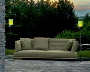 Zwei Akku-Windlichter auf hohen Gestellen flankieren ein grünes Sofa im Freien, umgeben von Natur bei Sonnenuntergang. Perspektive: Frontalansicht.