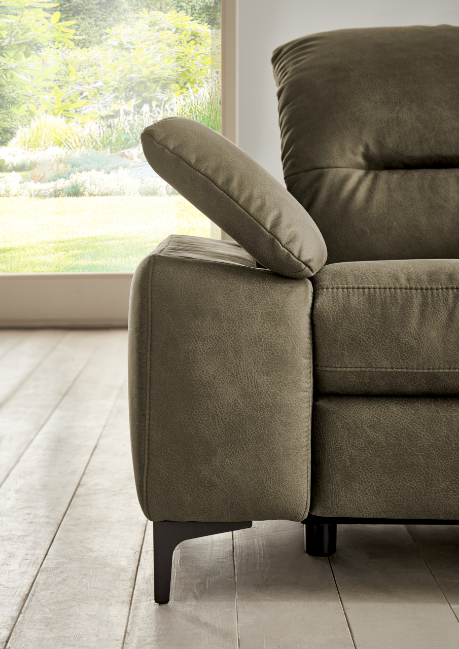 Nahaufnahme eines modernen Longchairs in Olivgrün mit verstellbarer Armlehne, seitliche Perspektive, im Hintergrund ein Fenster mit Blick ins Grüne.