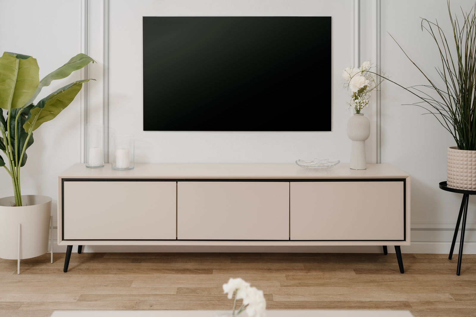 Modernes TV-Lowboard in heller Holzoptik mit schwarzen Akzenten, frontal betrachtet, dekoriert mit Pflanzen und Kerzen.