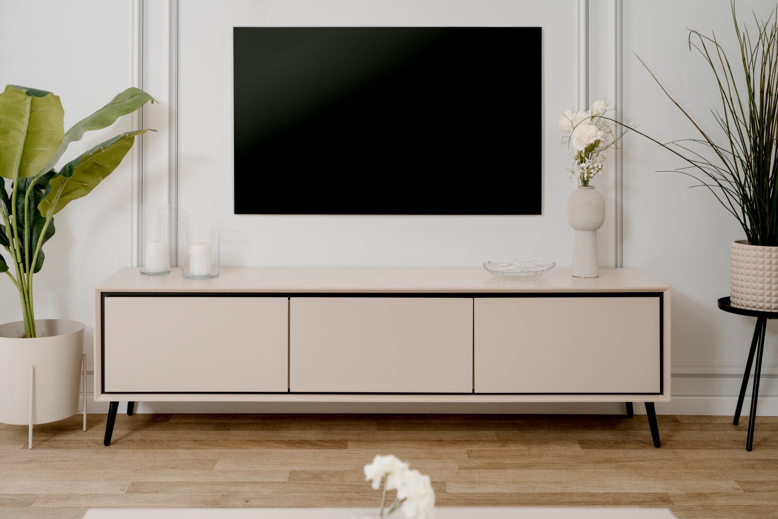 Modernes TV-Lowboard in heller Holzoptik mit schwarzen Akzenten, frontal betrachtet, dekoriert mit Pflanzen und Kerzen.