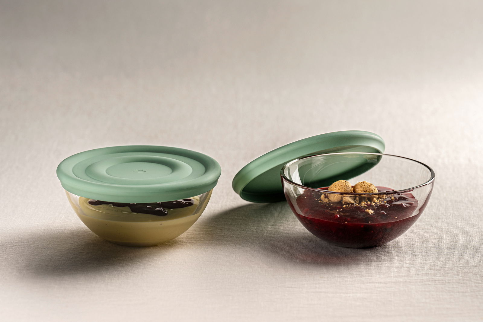 Zwei Glasschalen mit Salbei-farbenen Silikondeckeln, eine Schale mit Pudding und Schokoladensauce, die andere mit rotem Kompott und Keksen, seitliche Perspektive.