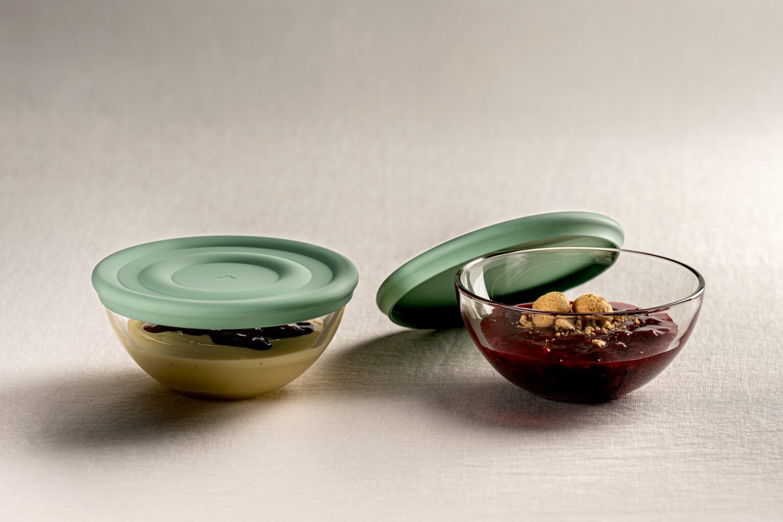 Leonardo Silikondeckel 14 salbei CUCINA Zwei Glasschalen mit Salbei-farbenen Silikondeckeln, eine Schale mit Pudding und Schokoladensauce, die andere mit rotem Kompott und Keksen, seitliche Perspektive.