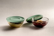 Zwei Glasschalen mit Salbei-farbenen Silikondeckeln, eine Schale mit Pudding und Schokoladensauce, die andere mit rotem Kompott und Keksen, seitliche Perspektive.
