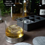 Nahaufnahme eines Whiskyglases mit einem großen Eiswürfel, im Hintergrund eine Eiswürfelform. Perspektive von vorne.