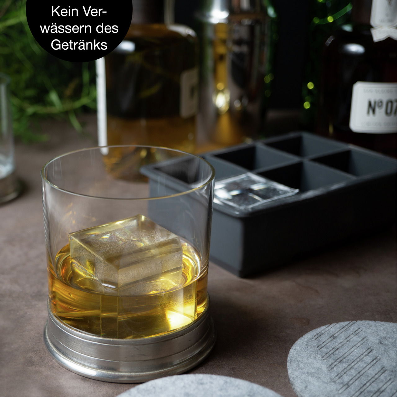 Nahaufnahme eines Whiskyglases mit einem großen Eiswürfel, im Hintergrund eine Eiswürfelform. Perspektive von vorne.