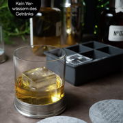 Nahaufnahme eines Whiskyglases mit einem großen Eiswürfel, im Hintergrund eine Eiswürfelform. Perspektive von vorne.