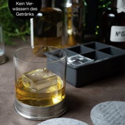 Nahaufnahme eines Whiskyglases mit einem großen Eiswürfel, im Hintergrund eine Eiswürfelform. Perspektive von vorne.