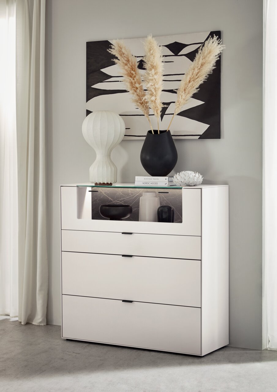 Frontale Ansicht eines modernen weißen Highboards mit drei Schubladen und einem offenen Fach, dekoriert mit einer Vase und Büchern.