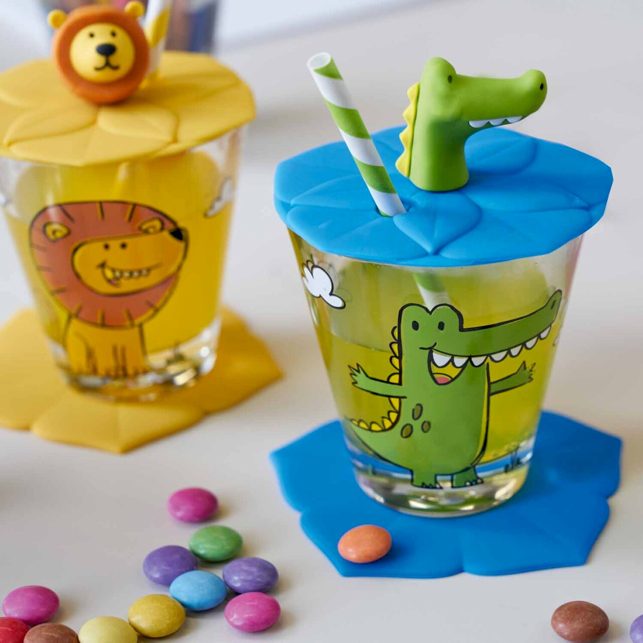 Kinderservies met krokodil- en leeuwenmotief, bestaande uit twee glazen met kleurrijke deksels en rietjes, gefotografeerd vanuit een schuin bovenaanzicht.