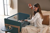 Frau sitzt auf einem Bett und schaut auf ein blaues Anstellbett mit einem Baby darin, seitliche Perspektive.