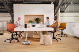 Drei Personen sitzen an einem modernen Konferenztisch auf farbigen Drehstühlen in einem hellen Büro mit Pflanzen im Hintergrund. Perspektive: Frontalansicht.