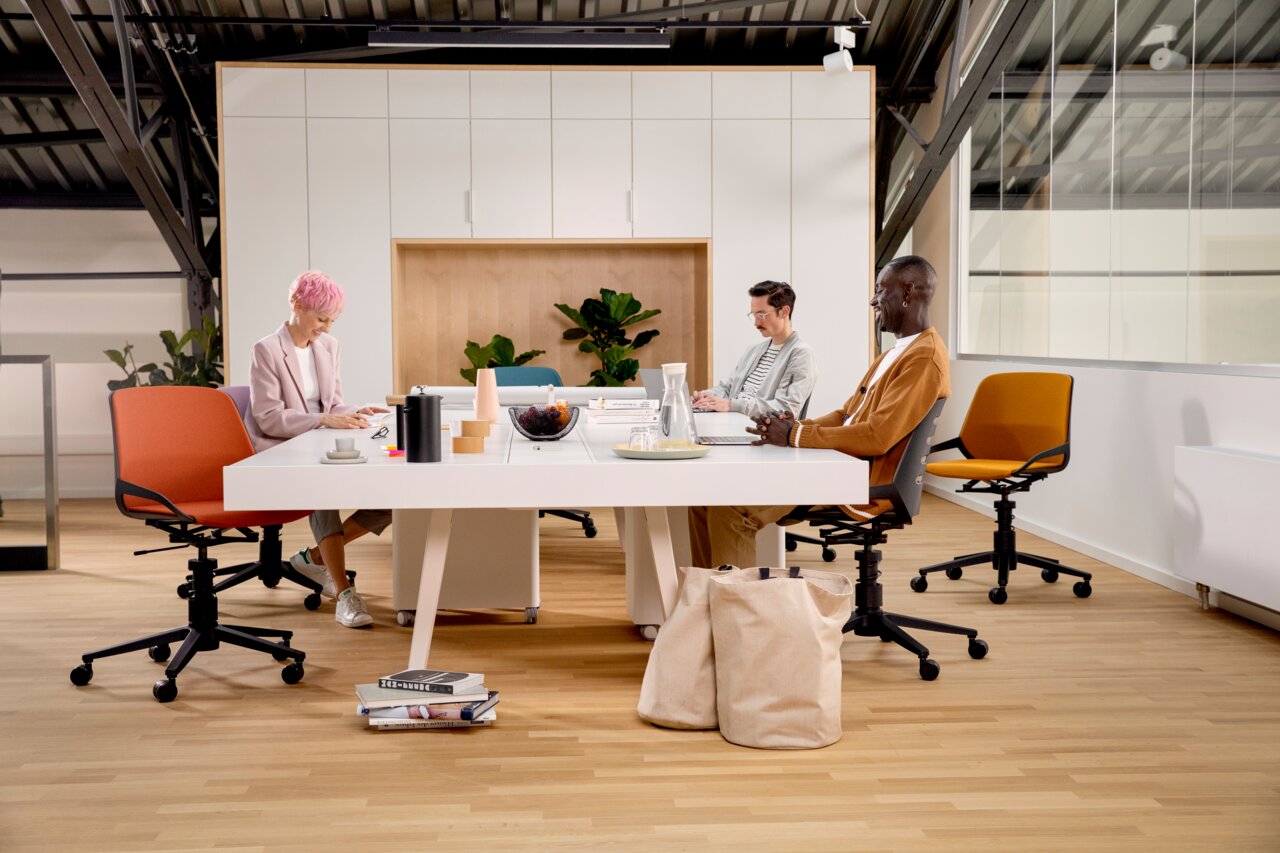 Drei Personen sitzen an einem modernen Konferenztisch auf farbigen Drehstühlen in einem hellen Büro mit Pflanzen im Hintergrund. Perspektive: Frontalansicht.